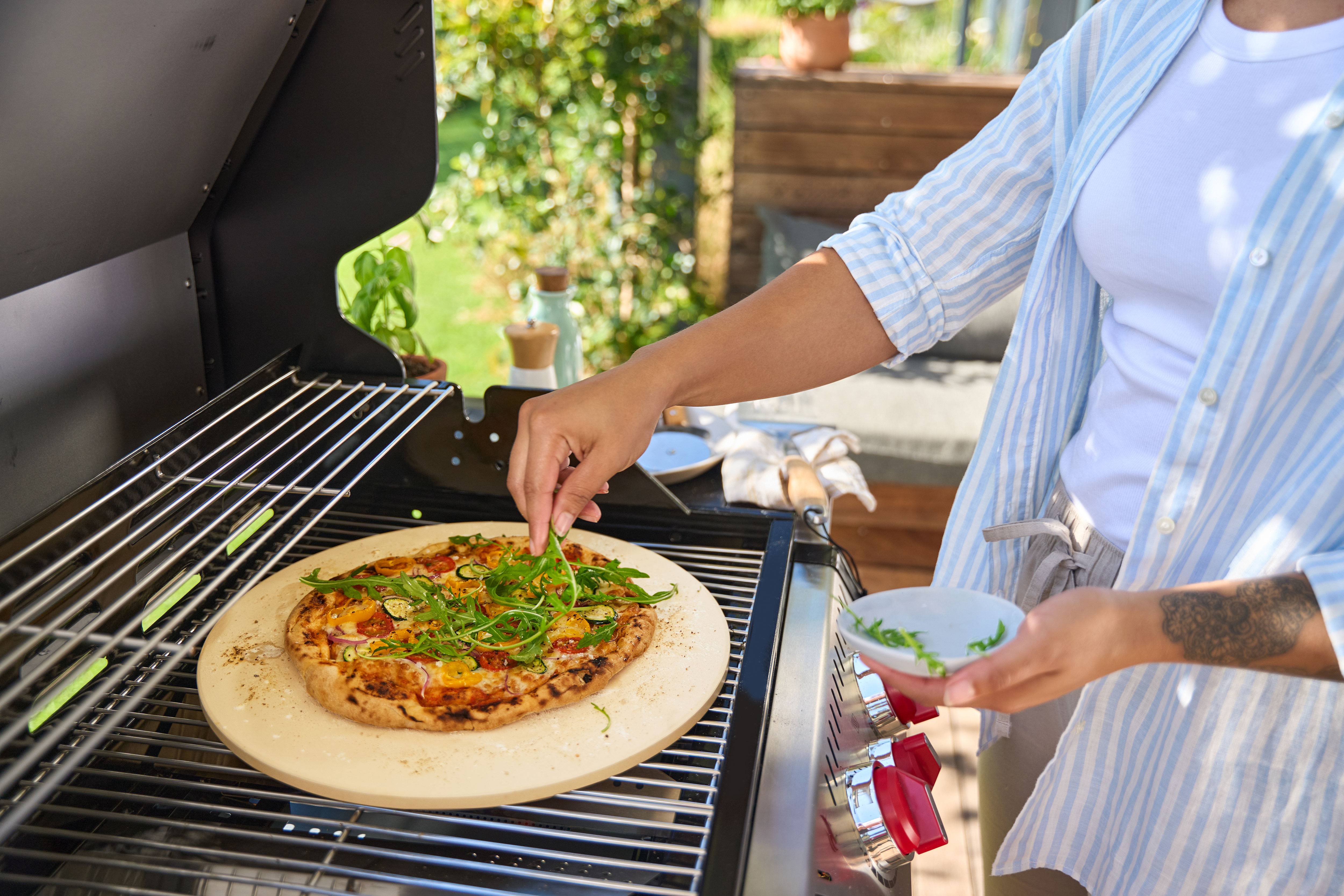 Osoba přidávající rukolu na pizzu na pizza kameni v plynovém grilu.