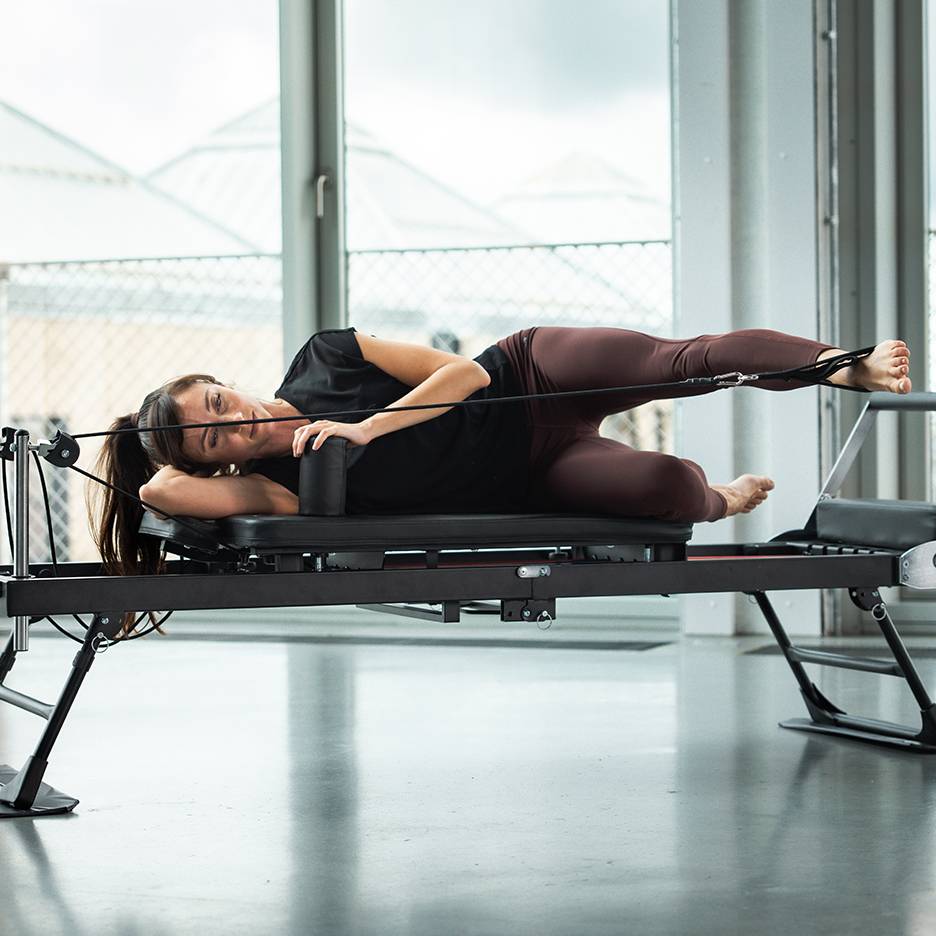 Žena cvičí Pilates na reformer stroji, ležící na boku s popruhy na nohou.