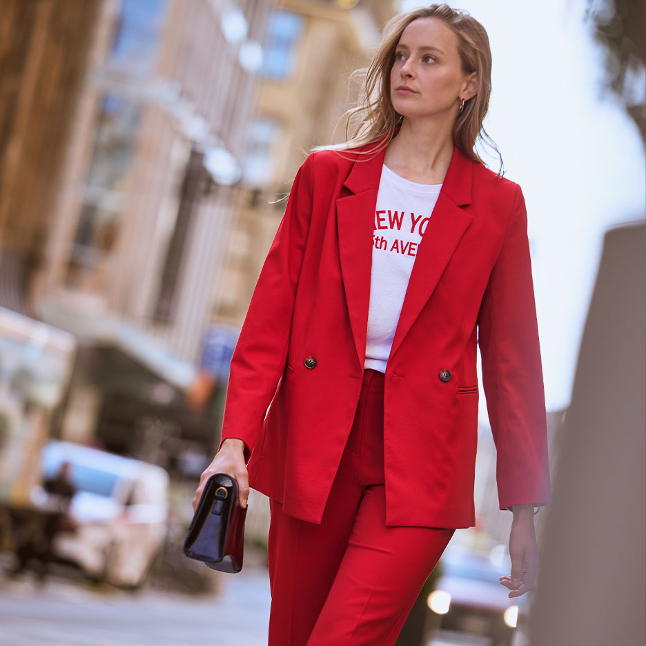 Žena v červeném kalhotovém kostýmu a bílém tričku s textem 'NEW YORK 5th AVE', drží černou kabelku.