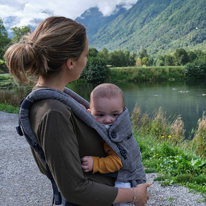 Matka nese své dítě v šedém ergonomickém nosítku na procházce u jezera.