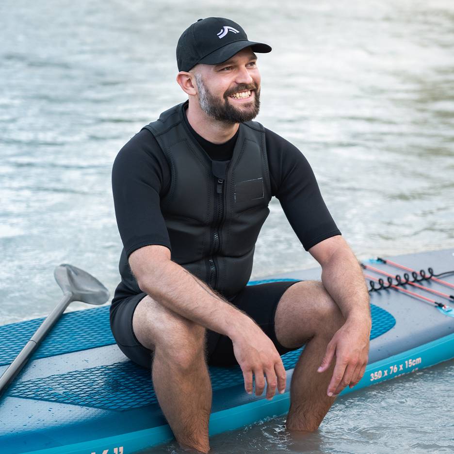 Usmívající se muž v záchranné vestě a čepici, sedící na paddleboardu ve vodě.