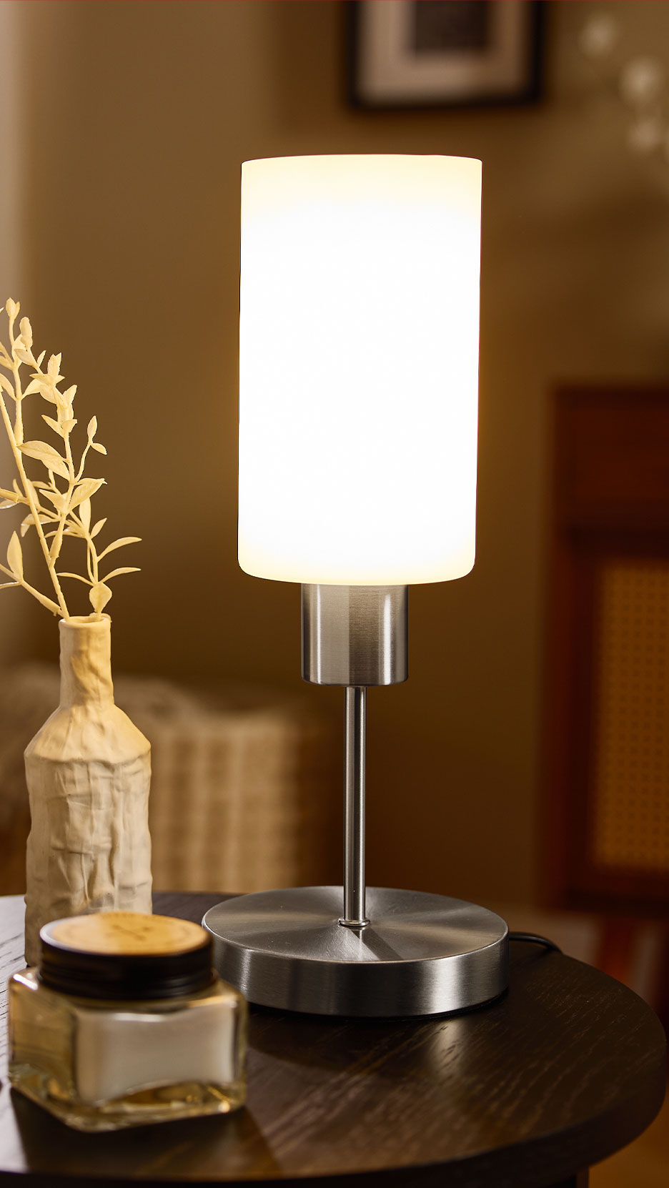 Moderní stolní lampa s bílým válcovým stínítkem a podstavcem z broušeného kovu na tmavém dřevěném stole.