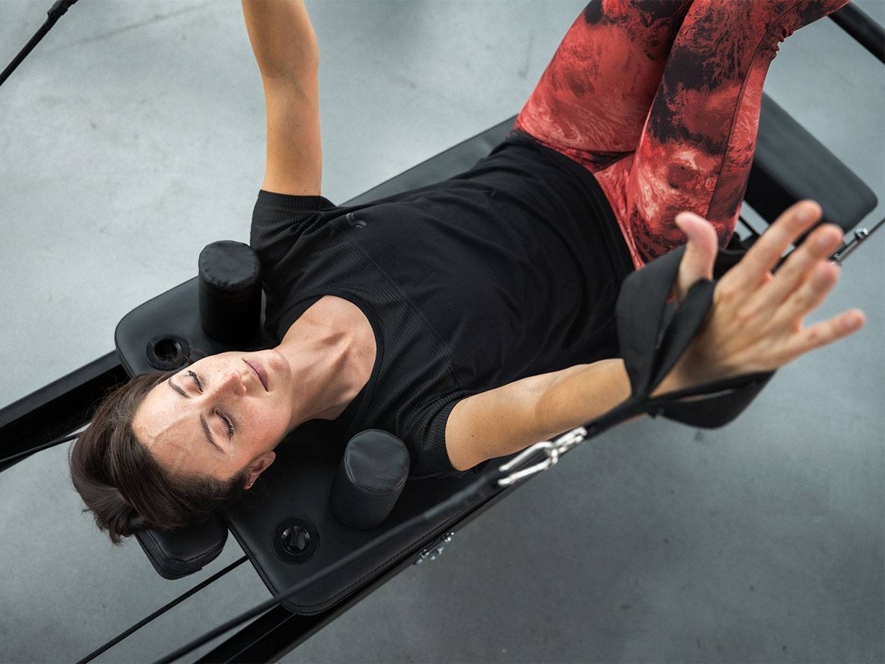 Žena ve sportovním oblečení leží na Pilates stroji a cvičí.