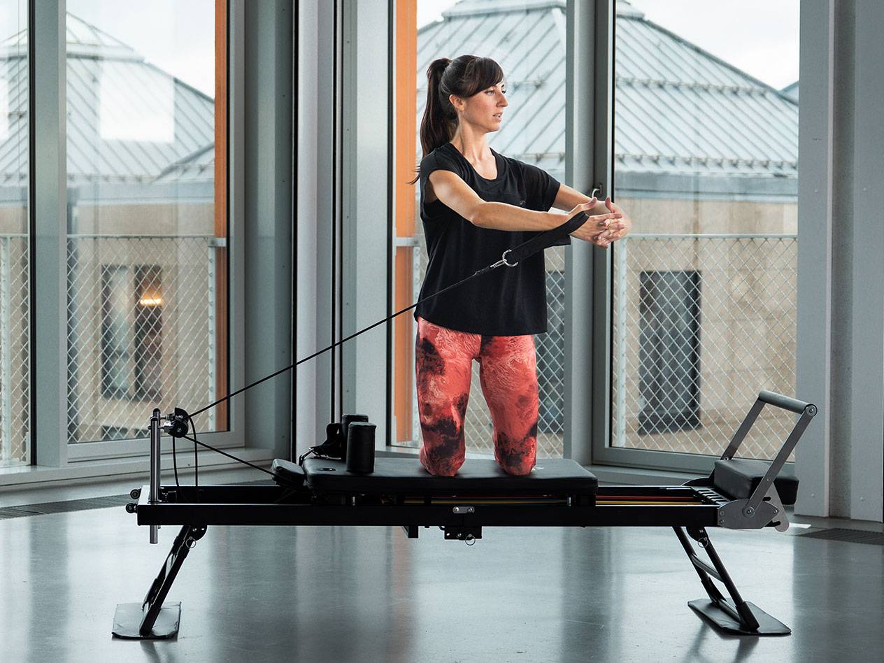 Žena cvičí Pilates na černém reformer stroji ve světlé místnosti.
