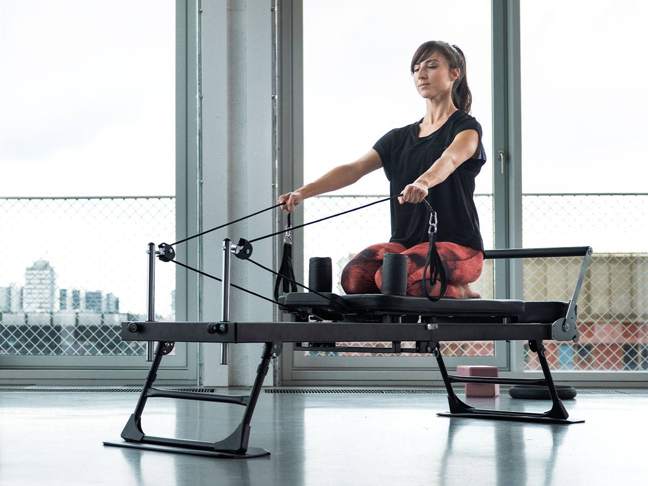 Žena cvičící Pilates na reformer stroji s pozadím města.