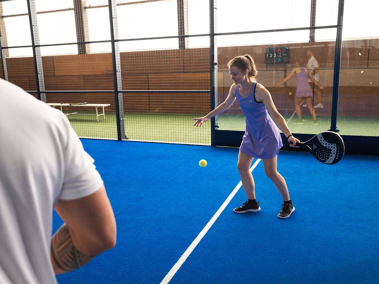Žena hraje padel, drží raketu a dívá se na míč na modrém kurtu.