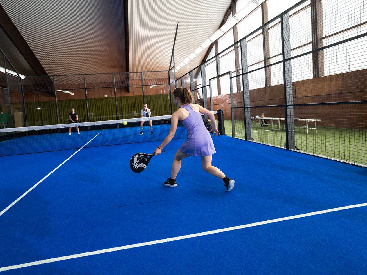 Žena hraje padel na modrém kurtu, s dalšími hráči v pozadí.