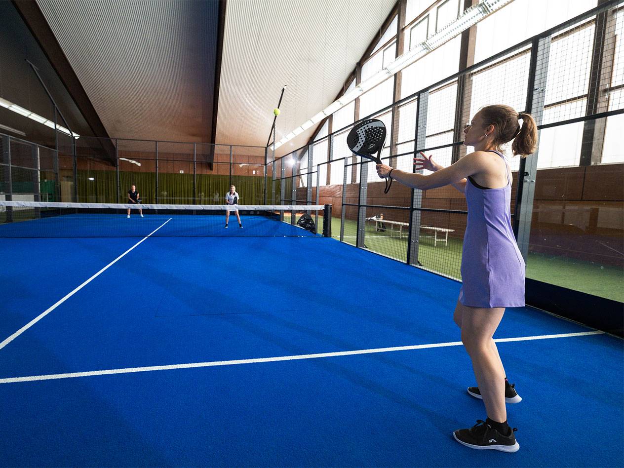 Žena ve fialových šatech hraje padel na modrém kurtu, ostatní hráči v pozadí.