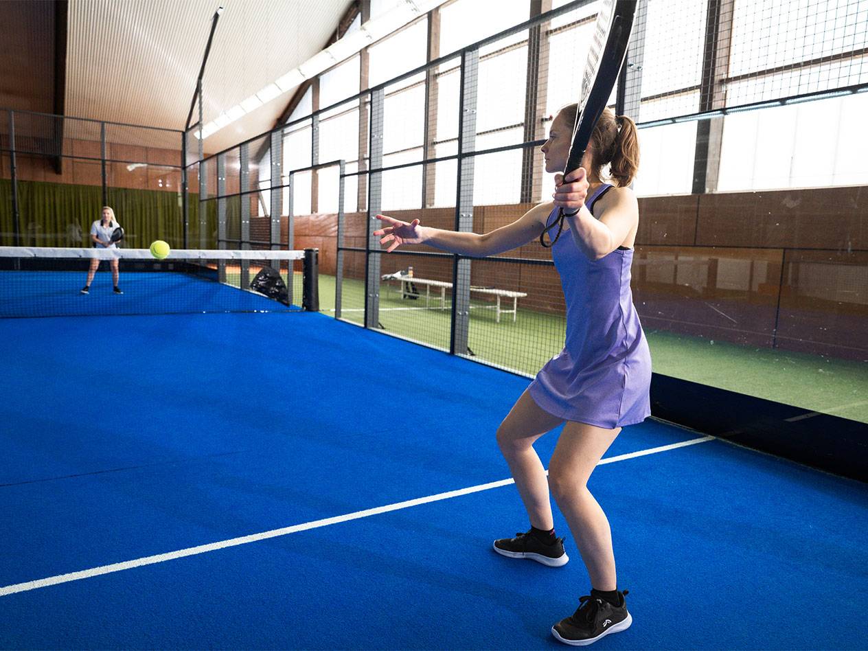 Žena ve fialových sportovních šatech hraje padel na modrém kurtu.