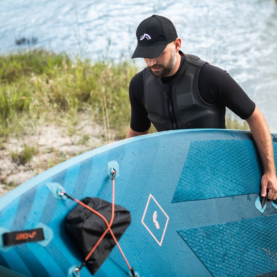Muž v záchranné vestě a čepici drží modrý paddleboard u vody.