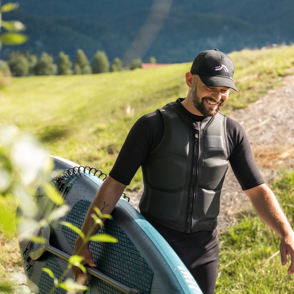 Muž v černém neoprenu a záchranné vestě nese paddleboard s pádlem.
