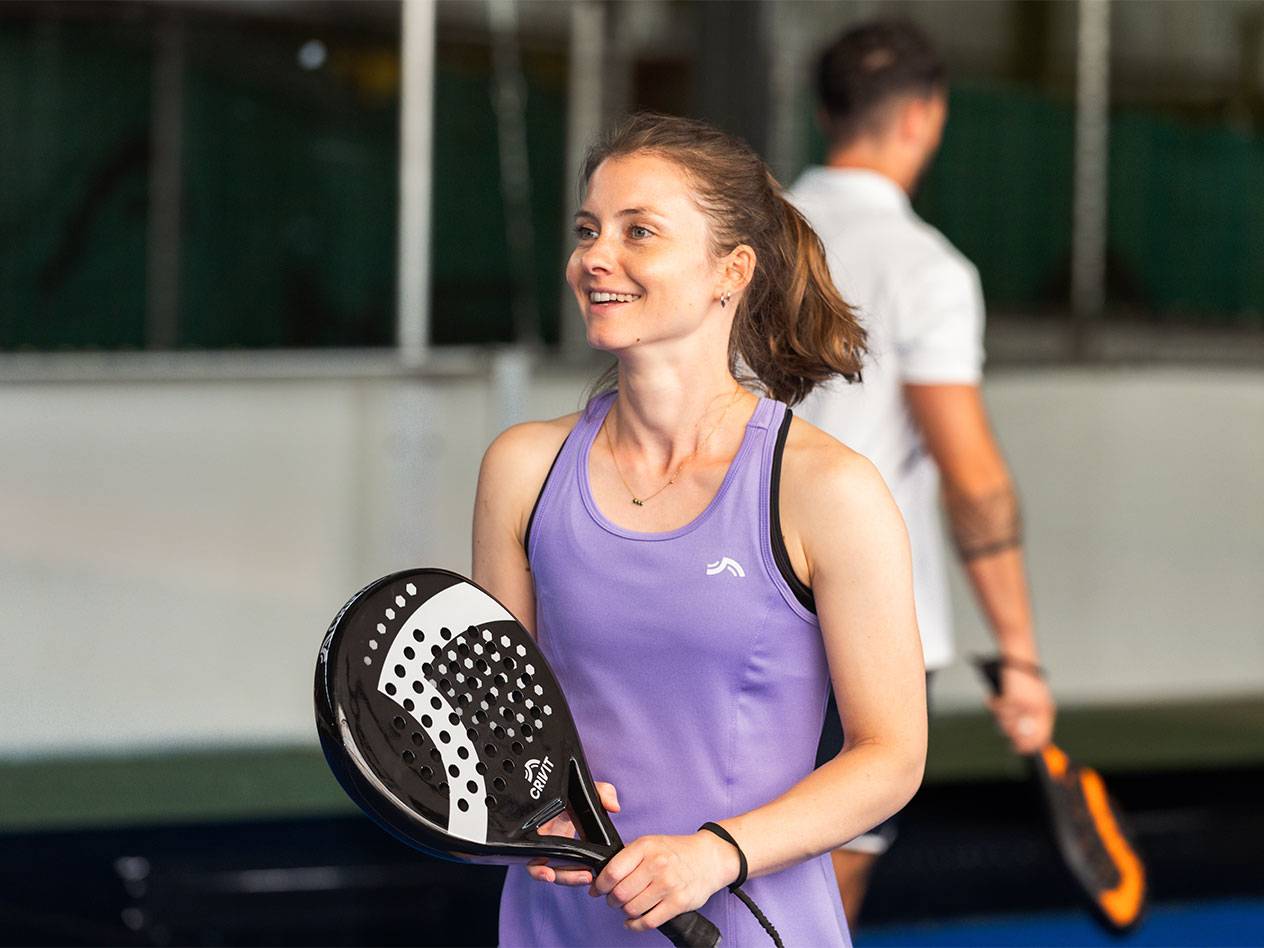 Žena ve fialových sportovních šatech drží padelovou raketu, muž v pozadí.