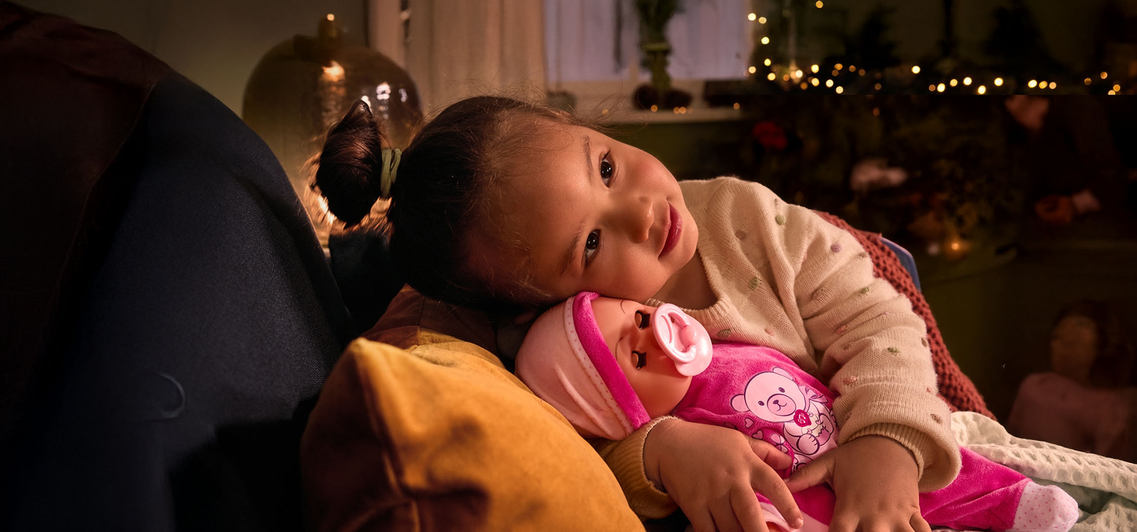 Holčička s panenkou a dudlíkem, útulně usazená na polštáři.