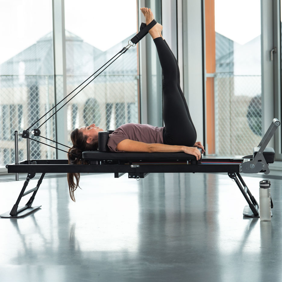 Žena cvičí Pilates na reformer stroji s nohama nahoře, ve světlé místnosti.