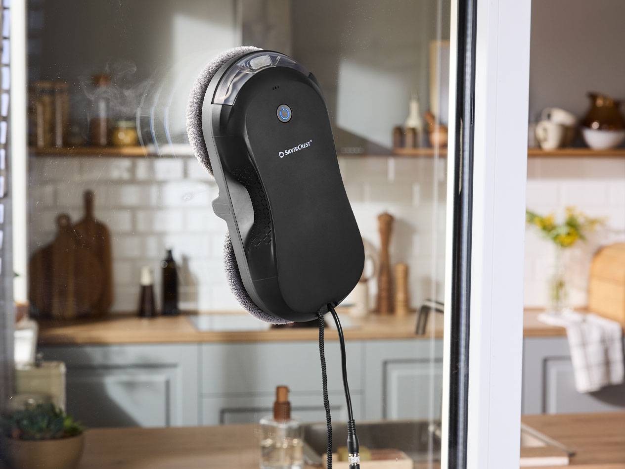 Robot na čištění oken Silvercrest na okenní tabuli s kuchyní v pozadí.