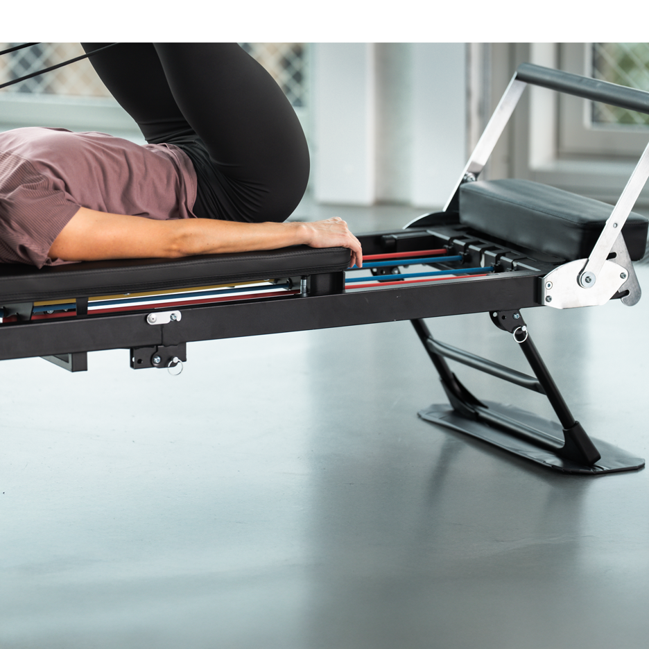 Žena cvičící na černém Pilates stroji s barevnými odporovými pásy.