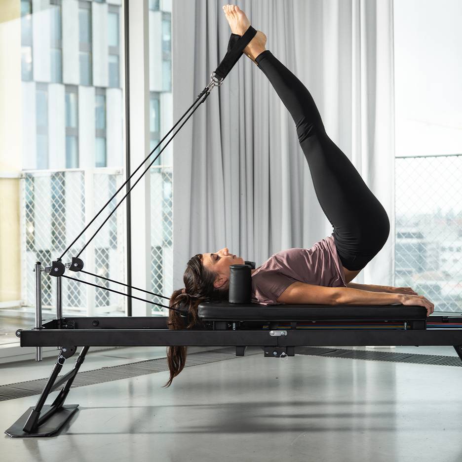 Žena cvičící Pilates na černém stroji s nohama nahoře.