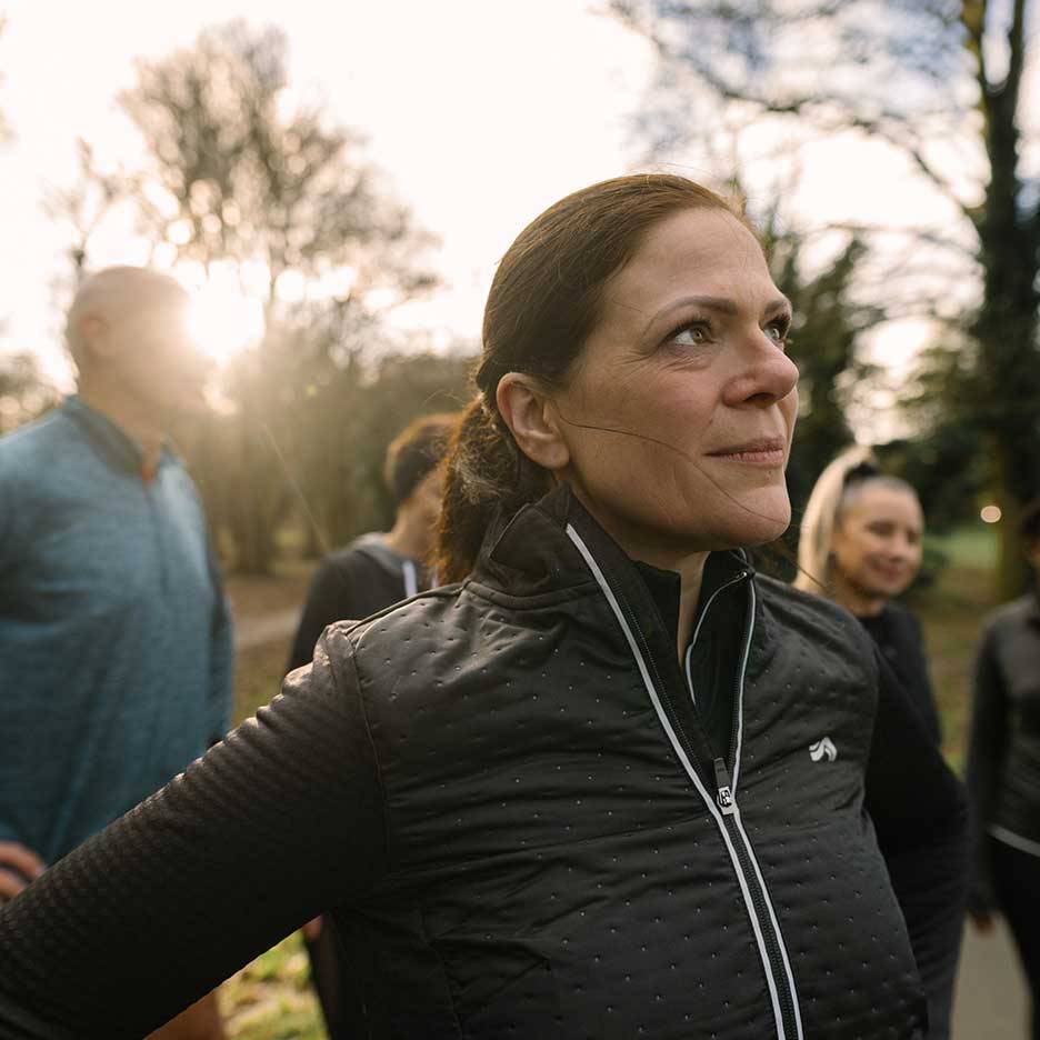 Žena v černé sportovní bundě se dívá nahoru, v pozadí skupina lidí.
