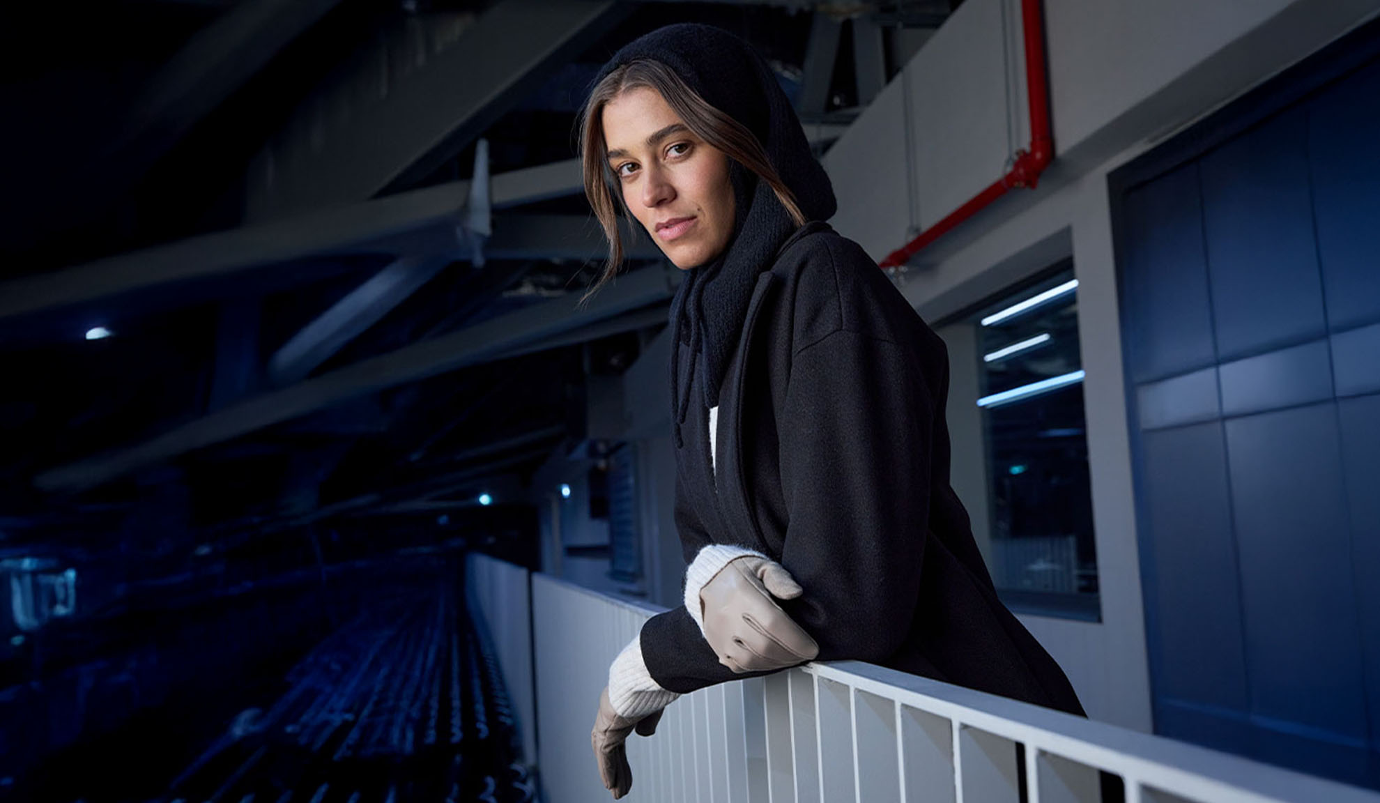 Žena v černém kabátě a šátku s kapucí, opírající se o zábradlí v moderním prostředí.