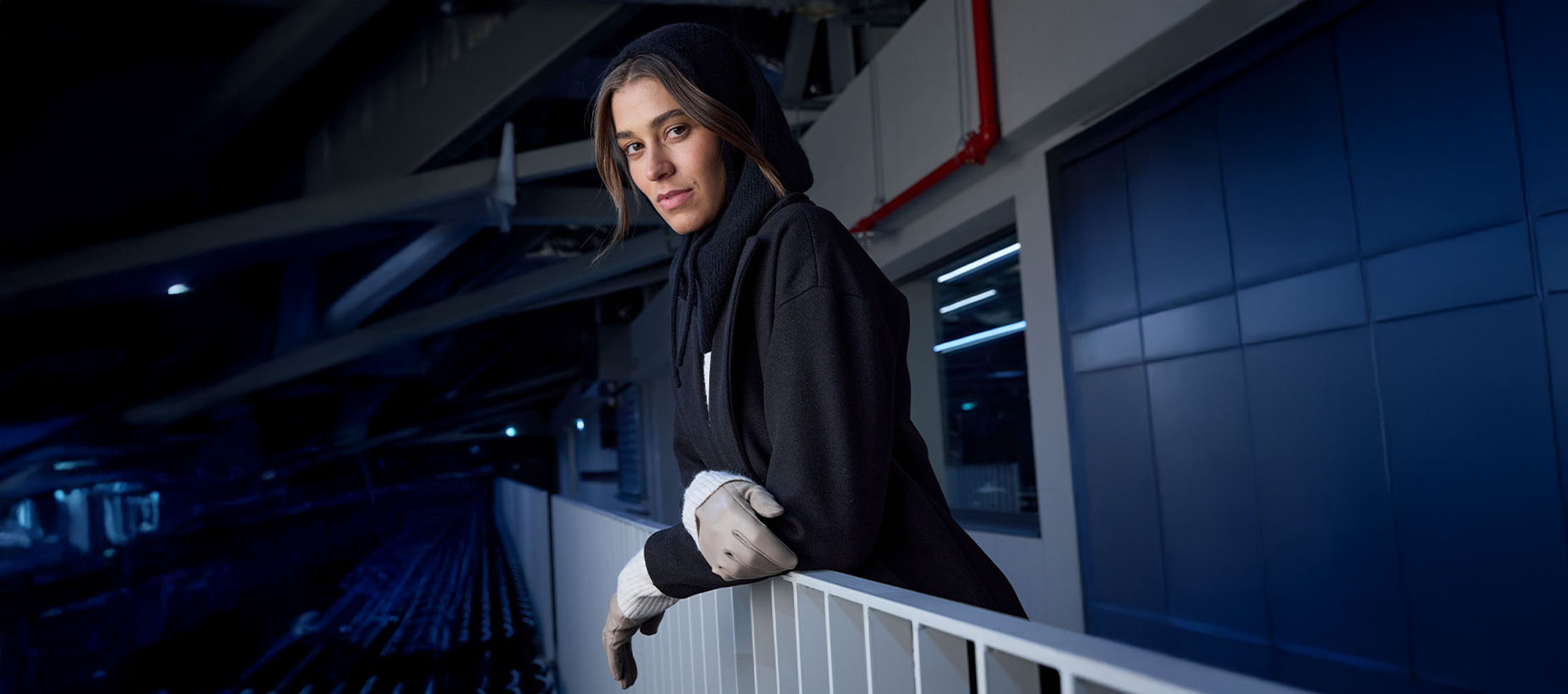 Žena v černém kabátě a rukavicích opřená o zábradlí v městském prostředí.