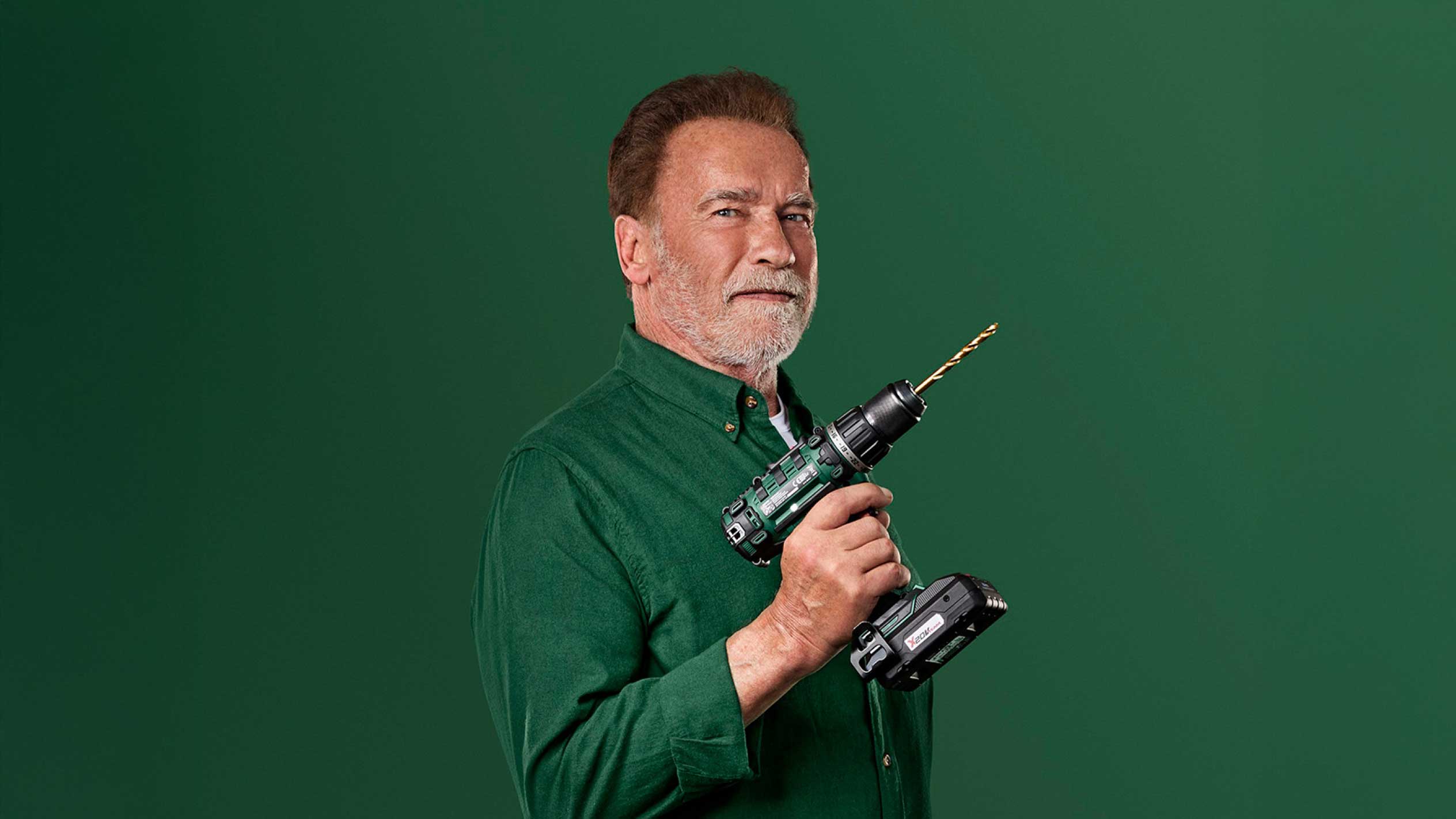 Arnold Schwarzenegger drží zelenou akumulátorovou vrtačku se zlatým vrtákem.