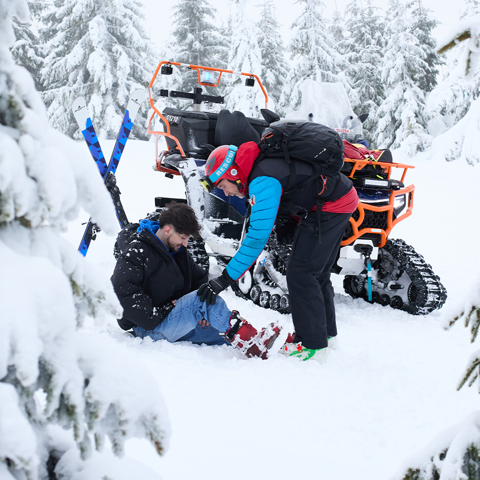Záchranář pomáhá zraněnému lyžaři v zasněžené krajině s terénním vozidlem v pozadí.