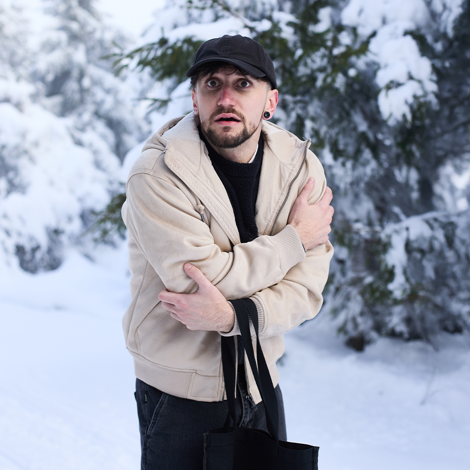 Muž v béžové bundě a černé čepici se třese zimou ve sněhem pokrytém lese.