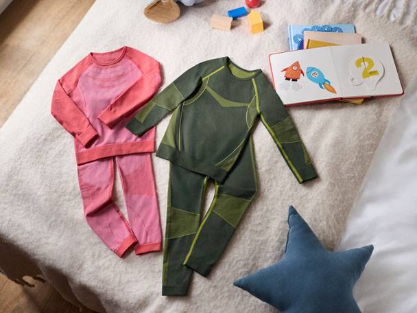 Dvě sady dětského termoprádla (růžové a zelené) a kniha na posteli.
