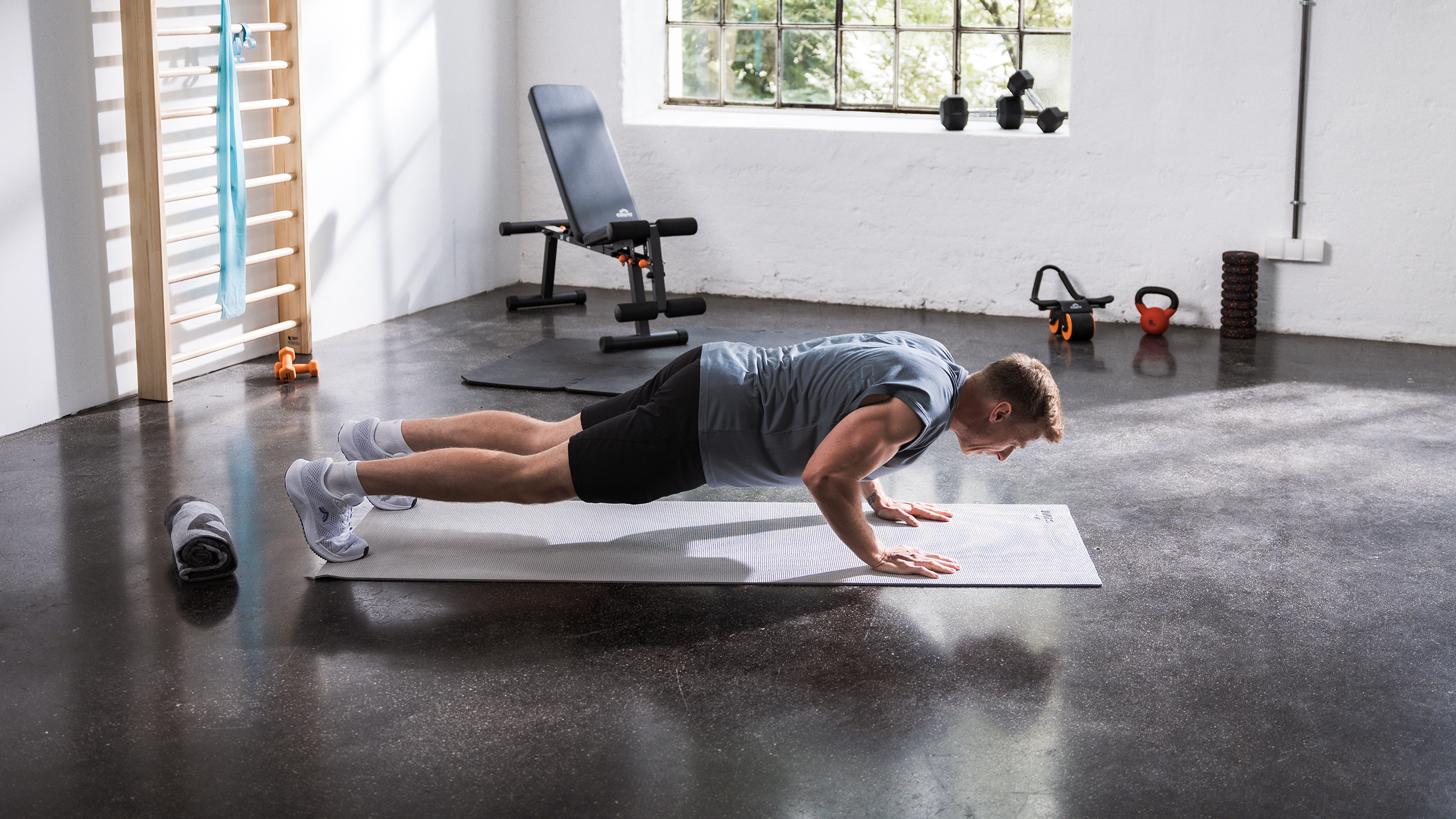 Muž dělá kliky na podložce v domácí posilovně s různým fitness vybavením.