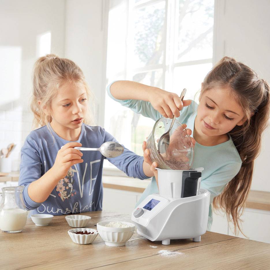 Dvě dívky připravují těsto v kuchyňském robotu, s miskami ingrediencí na stole.