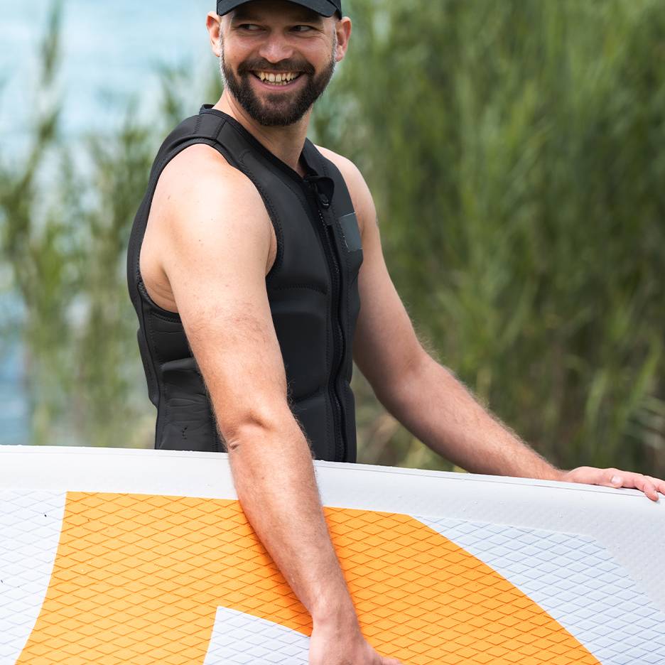 Usmívající se muž v černé záchranné vestě drží paddleboard.