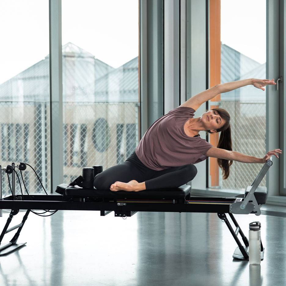 Žena cvičící pilates na reformer stroji, protahující se do strany.