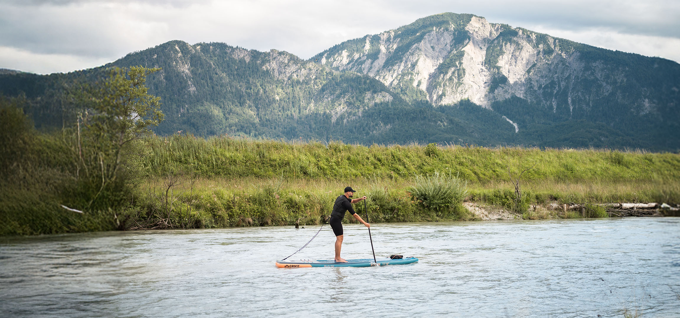 Muž na paddleboardu na řece s horami a bujnou vegetací v pozadí.