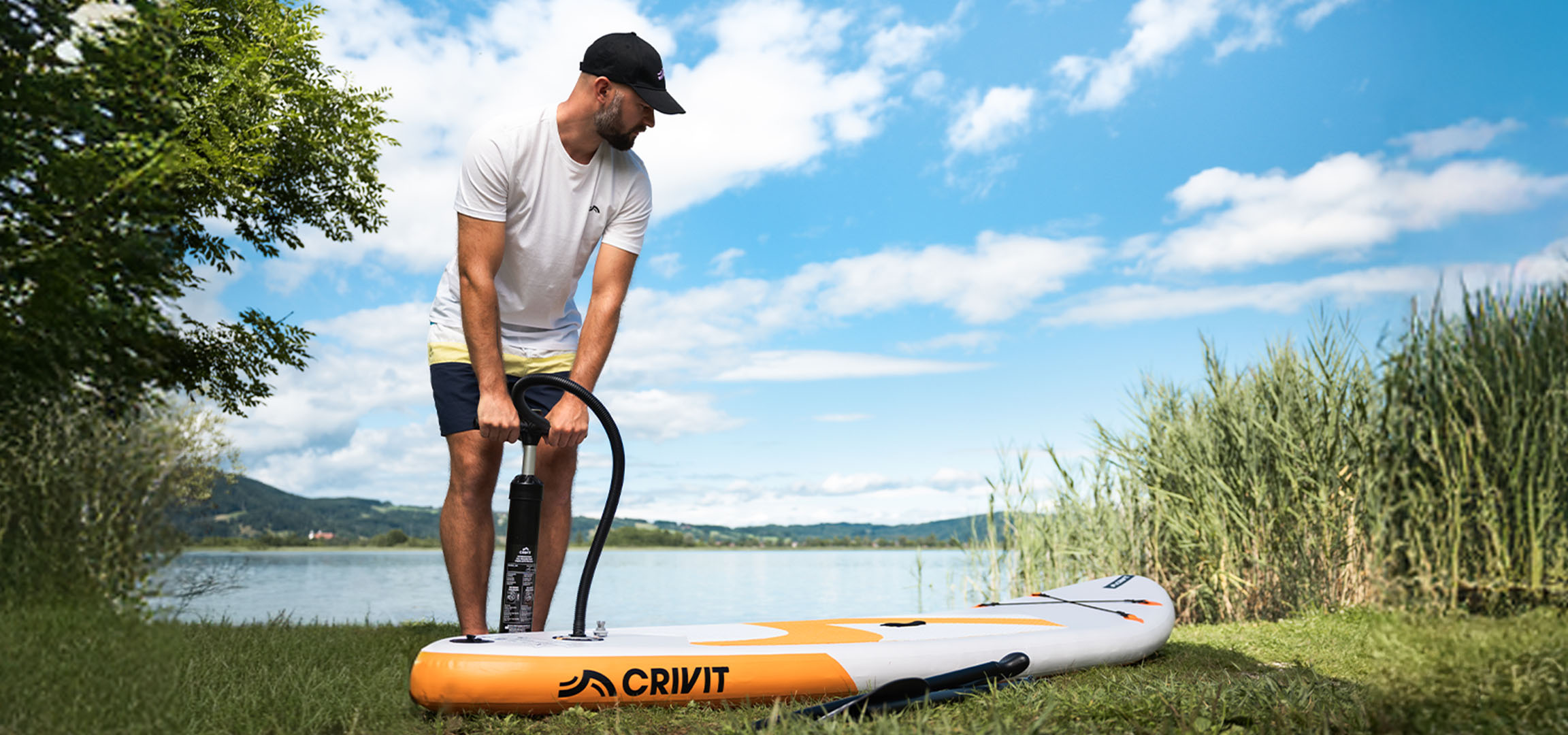 Muž nafukuje paddleboard CRIVIT u jezera na zelené trávě.