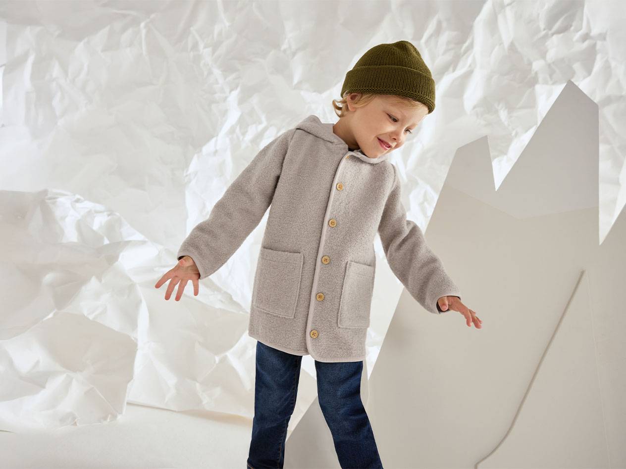 Chlapec v šedém vlněném kabátě a zelené čepici, usmívá se na pozadí zmačkaného papíru.