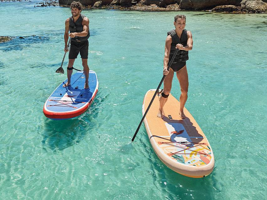 Dva lidé na paddleboardech s pádly a záchrannými vestami v tyrkysové vodě.