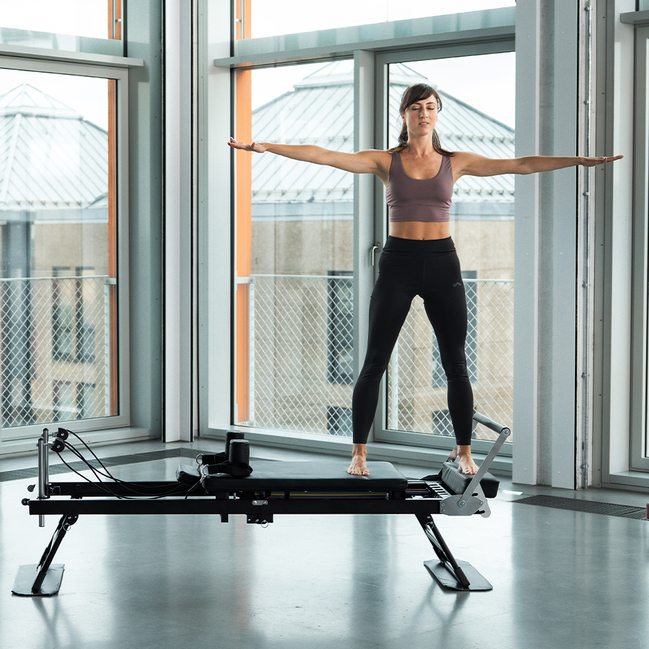 Žena cvičící Pilates na reformeru s rozpaženýma rukama.