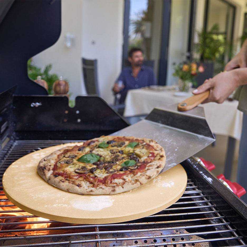 Pizza na pizza kameni na grilu, s lopatkou na pizzu a mužem v pozadí.