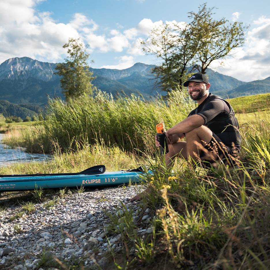 Usmívající se muž v kšiltovce a záchranné vestě, sedící u modrého paddleboardu u řeky.