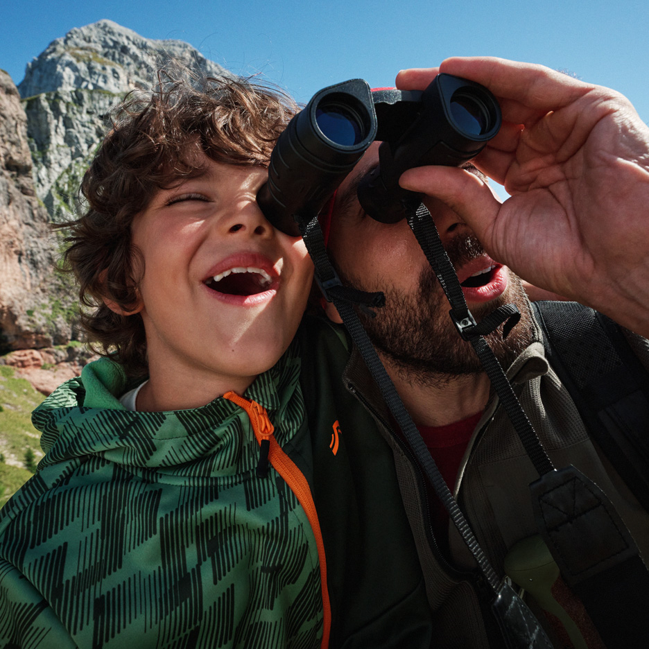 Otec a syn prozkoumávají hory s dalekohledem, v outdoorových bundách.