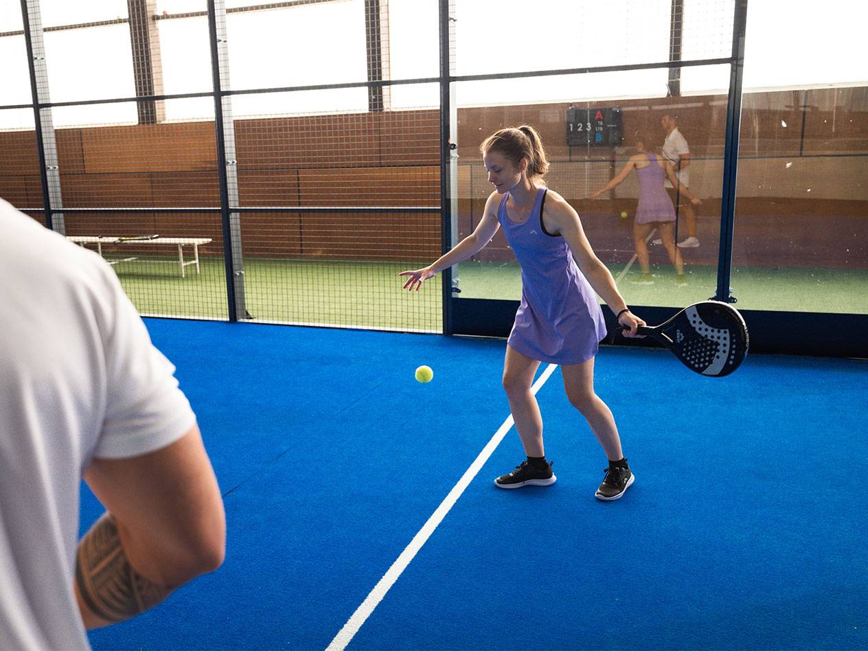 Žena ve fialových sportovních šatech hraje padel s raketou a míčkem.