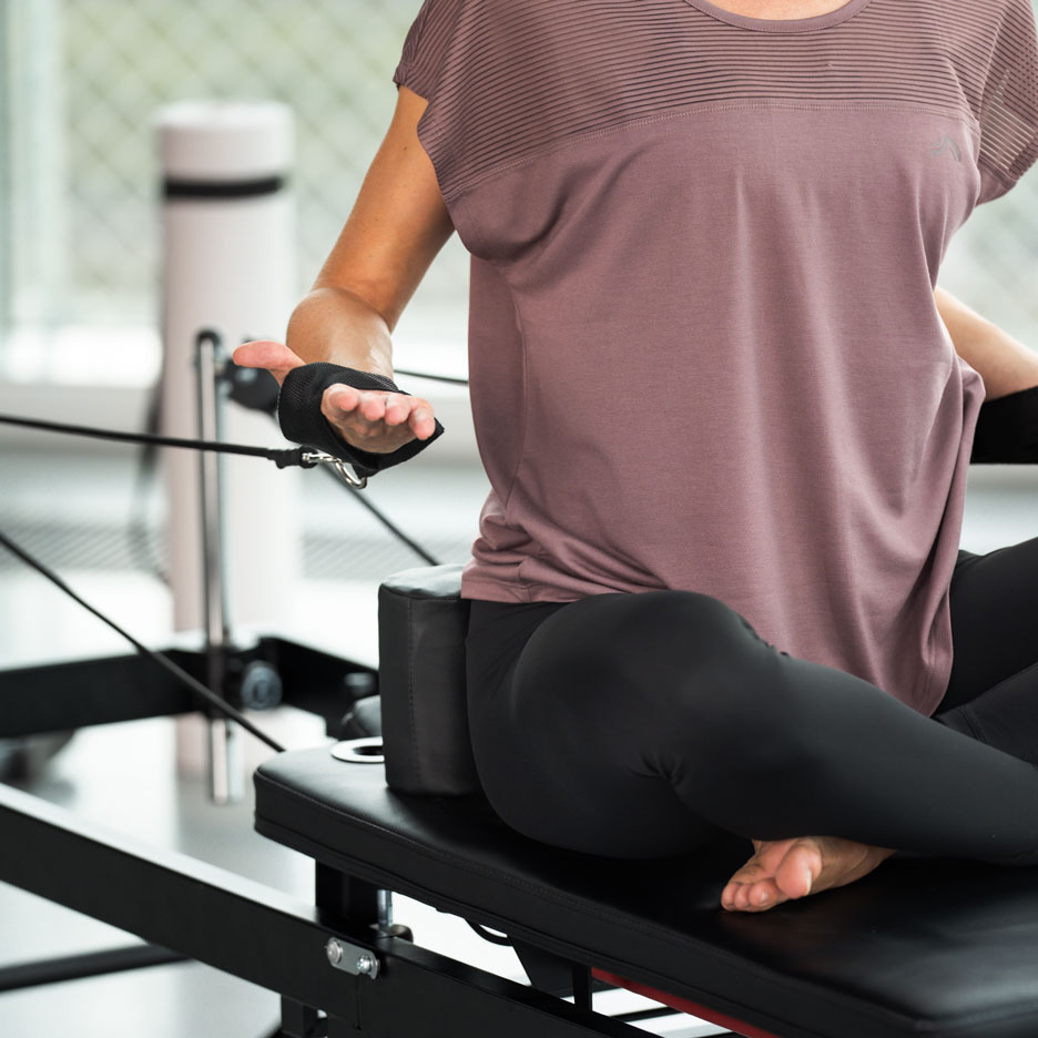 Žena cvičící Pilates na reformer stroji, v černých legínách a fialovém topu.