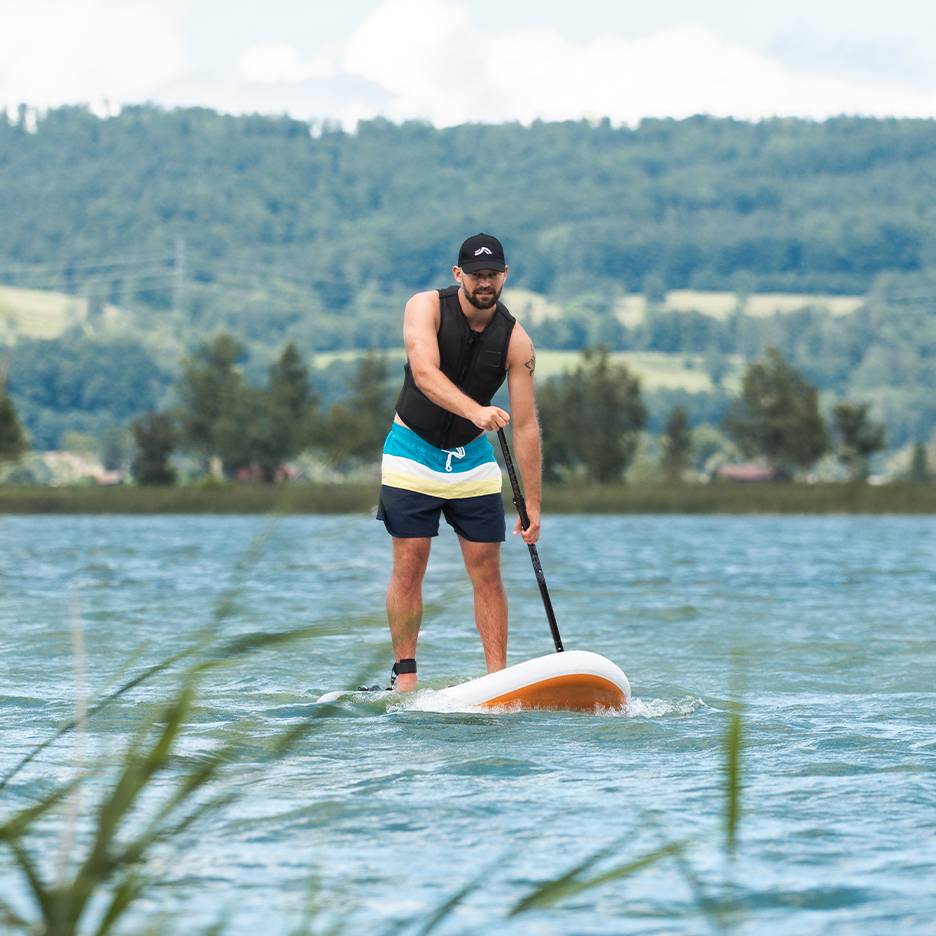 Muž na paddleboardu na jezeře, v záchranné vestě a plavkách.