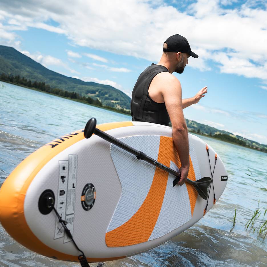 Muž v záchranné vestě a čepici drží nafukovací paddleboard v jezeře.