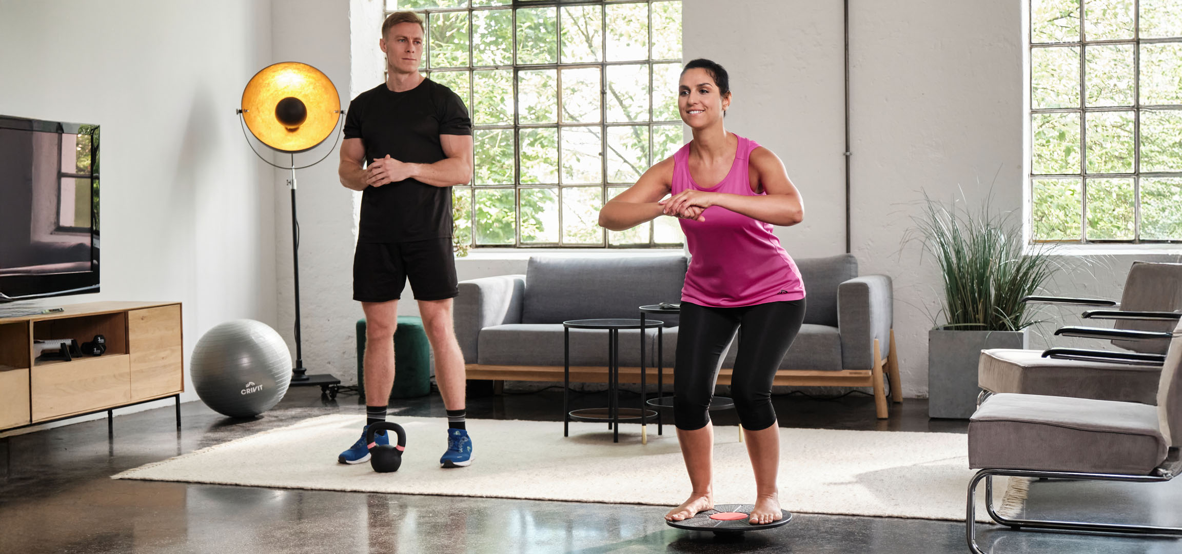 Žena a muž cvičí doma s balanční deskou a kettlebellem.