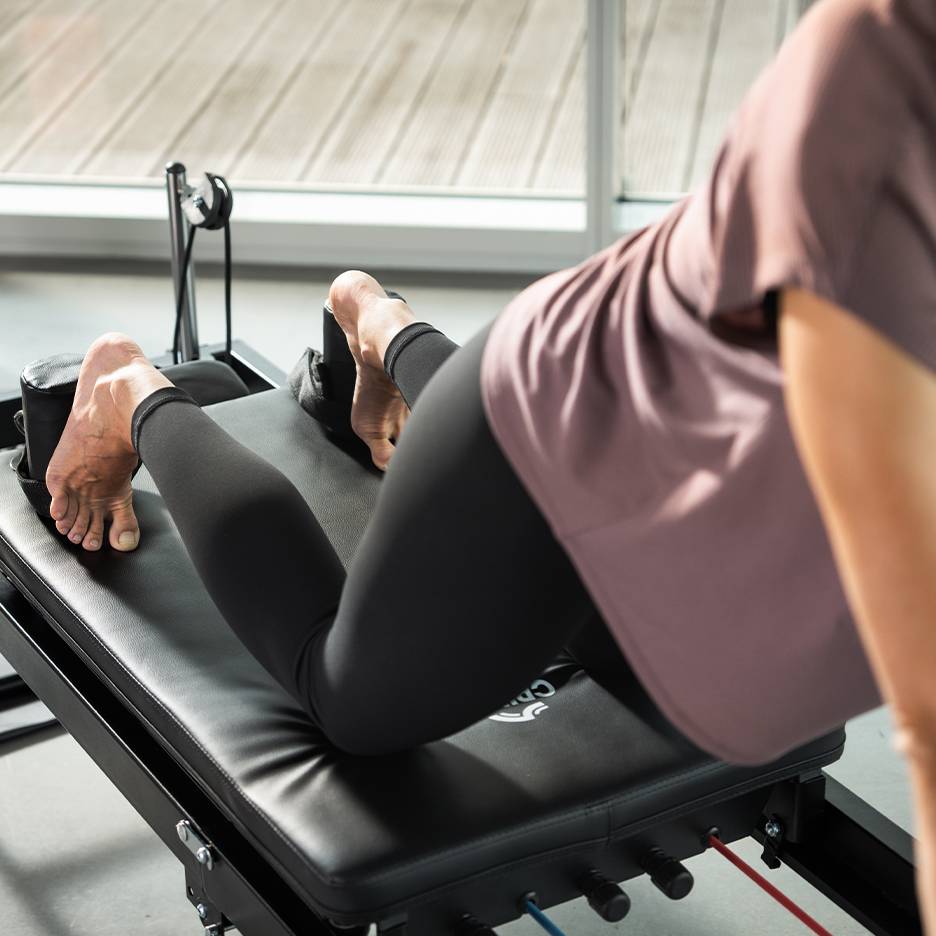 Žena cvičící na černém Pilates stroji s nohama v popruzích.