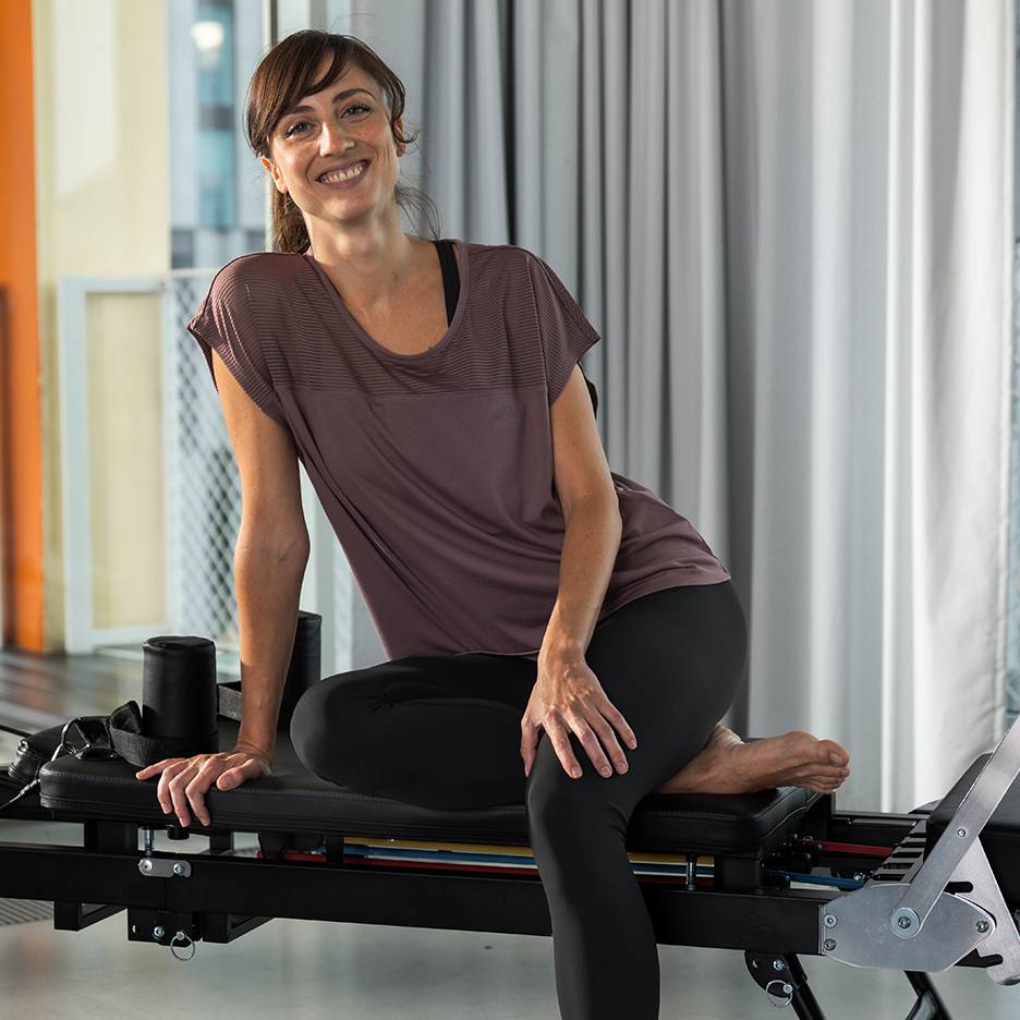 Žena ve sportovním oblečení se usmívá, sedí na stroji na pilates.