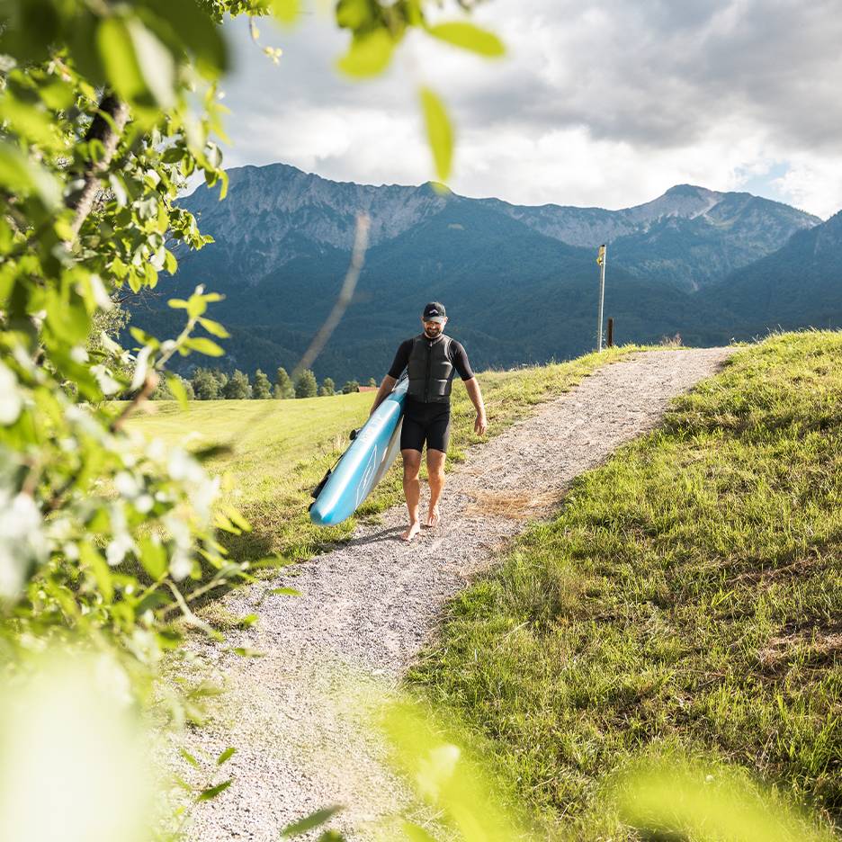 Muž v neoprenu a záchranné vestě nese paddleboard v horské krajině.