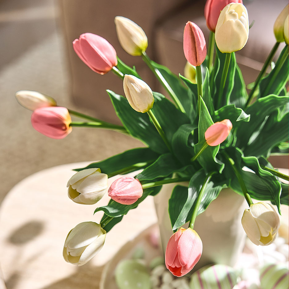 Růžové a bílé tulipány ve váze, s velikonočními vajíčky na podnosu v pozadí.