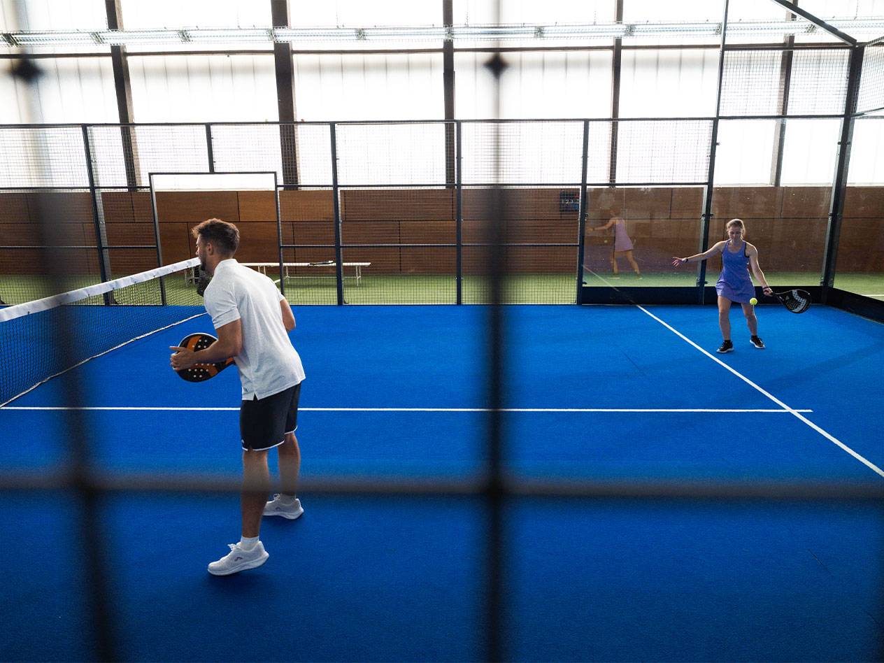 Muž a žena hrající padel na modrém kurtu s raketami a míčkem.